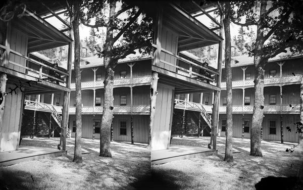 Courtyard at Cliff House, a resort hotel built shortly after 1872 when Devil's Lake was reached by rail. The house has a stone first floor, a balcony on each floor, and small gables on the roof. There is a man sitting on the second level.