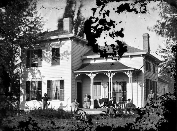 Some men are in a yard sitting and standing with scythe, rake and pitch fork. A woman sits on the front porch of a frame house with shutters, one closed on upper story, brackets and lightning rods. No doubt one of the men is Jerome Skaalen, and the woman his wife Rebecca.