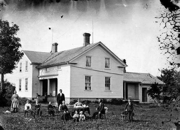 Family posed in yard around a table set for coffee. Behind is a large frame house with stone foundation, latticework at bottom of front porch, and lightning rods. A carriage sits at right and a boy is holding a dog on a chair.