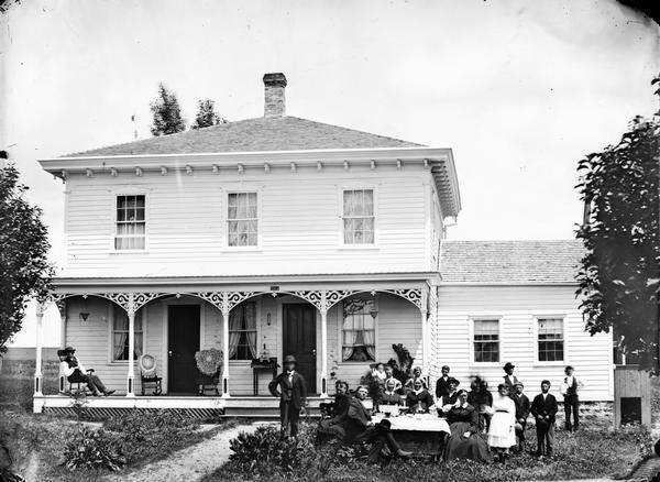 A large family is posed in the front yard around a table set with a white cloth and glassware, before a two-story bracket style house with a hip roof. The porch has carpenter's lace brackets, and on it are chairs displaying textile handicrafts, and a table with a bird cage. A man is sitting in a chair on the porch on the left.