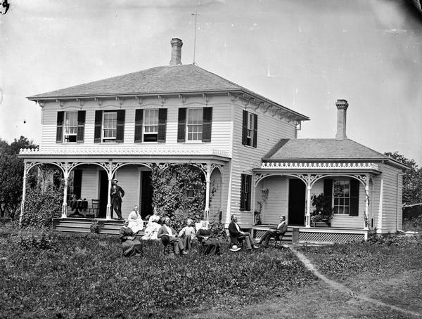 Large bracket house with shutters and man on porch; carpenter's lace in excess; family seated in yard in front of house. House also has latticework at foundation.