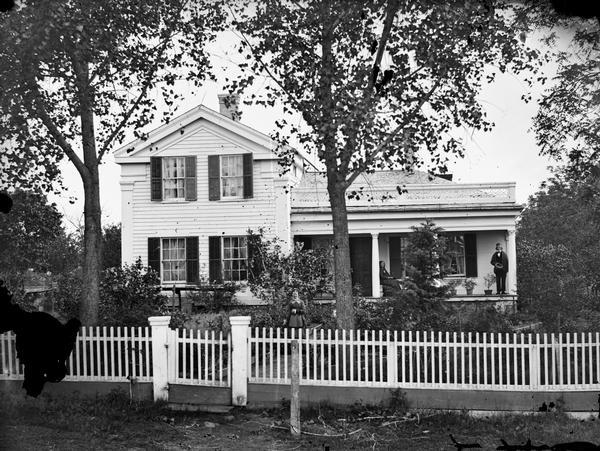 Greek revival frame house with Doric columns. A man is standing on the porch tipping his hat. A woman is sitting on the porch, and a woman is standing on a walkway in the foreground. There is a wood picket fence in front.