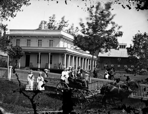 People in carriage in front of picket fence, wagons and people behind; large frame house with parapet roof, Corinthian porch columns, brackets at roof; carriage house is in background.