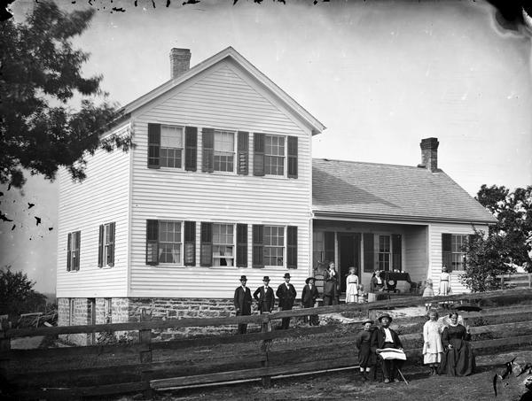 A family of fourteen in yard outside upright and wing frame house on stone foundation, with board fencing in the foreground.