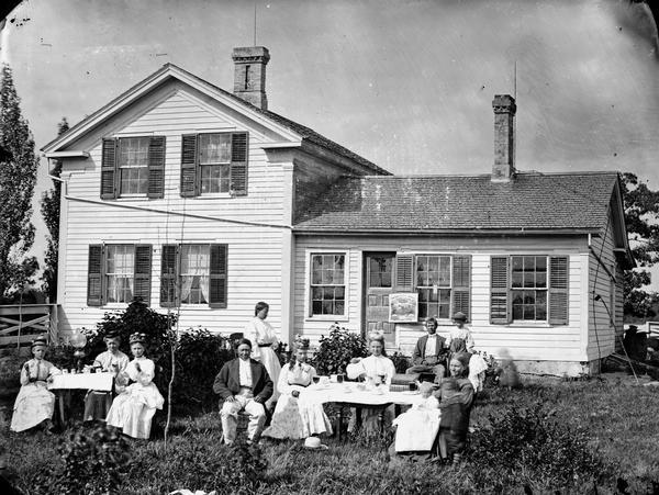 Family in front yard around two tables set for coffee, tea and perhaps some other beverage. Frame house in back has a sign for grass seed from Whitewater on front porch, and there is a lamp on one table in yard.