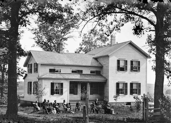 A family of twelve is seated in chairs in the yard of an upright and wing house.