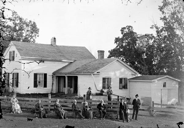 Family sitting in chairs in front of fence, with two men standing behind the fence near a well wearing top hats. A frame house with a porch is behind them.