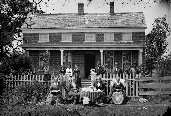 A family group is posing in yard. Part of the group is sitting around a table in front of a picket fence which encloses the brick Greek Revival house. Another group is standing behind the fence. One woman on the right is sitting and displaying a framed oval portrait, and the boy behind the fence on the left is holding an accordion.