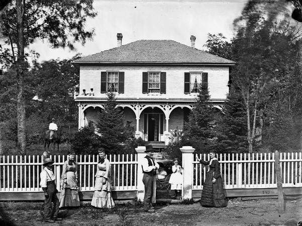 Family in front of picket fence, with girl and baby carriage behind gate, man on horse, brick house with hipped roof and shutters, and two boys on porch roof top in background.  This house is said to resemble the J. Hill house on Hubble Street in Black Earth, Wisconsin.