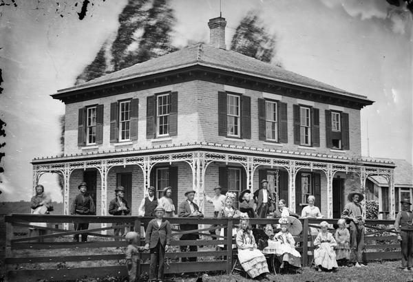 Christiana Township, Wis. Section 18. A group of twenty-one people in front of large brick house that has shutters, porch on at least two sides, trim around porch and hipped roof. Home and farm of Halvor Nerison Hauge (1830-1906).