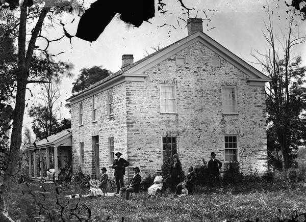 Family seated in yard of stone house, which has returned cornice characteristic of the Greek Revival style. There is a frame wing in the back.