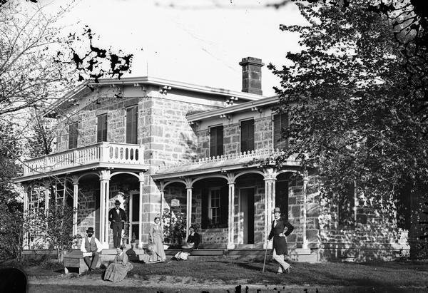 Harmon J. Hill and family, all dressed formally, posing in front of their home. This sandstone house was built in 1857 and later became part of the University Hill Farms acreage.