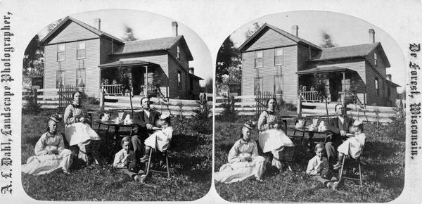 A husband, wife and three children are grouped around a table set for coffee. Behind them is a board fence and a frame house.
