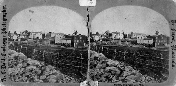 Probably one of the "Three different views of the village of Argyle, Wis." mentioned in Dahl's 1877 "Catalogue of Stereoscopic Views." A view of the town across a rustic fence toward a small stream.