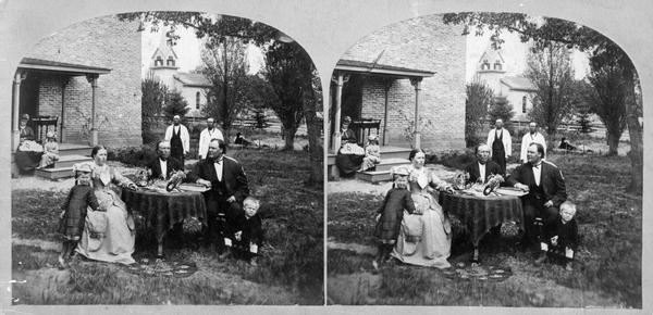 Mrs. Ingeborg Swenholt Homme sits at the left and her husband the Reverend Even Johnson Homme sits at the right, with their children, around a table in front of their home. The photograph was made at the meeting of the Eastern District Norwegian Synod, held at the Immanuel Norwegian Evangelical Lutheran Church (which can be seen in the distance).