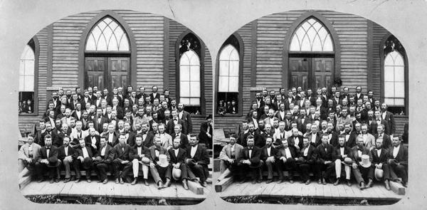 Stereograph of a group portrait of men assembled on the steps to a church door. "The Ministers" gathered at "The Norwegian Lutheran Synod held at Minneapolis, Minn., 1875," as described in Dahl's 1877 "Catalogue of Stereoscopic Views." Spectators are watching from the tops and bottoms of the windows on each side. A small sign on the left reads: "Washington Av. S."