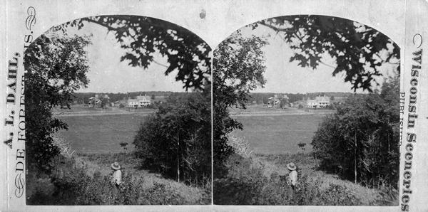 Allen E. Adsit is standing on a grassy hillside in the foreground.  In the distance is the Allen E. Adsit farm which includes a three-story frame house with a porch and several farm buildings.