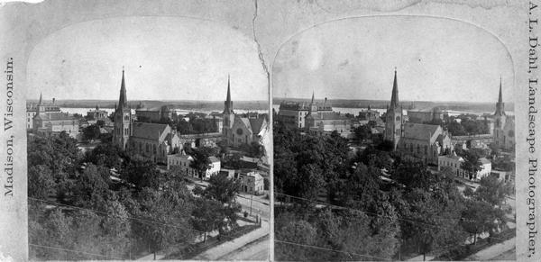 Stereograph looking south from the post office at the corner of Mifflin Street and Wisconsin Avenue across one corner of the Capitol Square toward Grace Episcopal Church (center), the Congregational Church (right), the Baptist Church (left) and the Park Hotel (far left). Lake Monona is in the background. Probably from the series "The Beauties of the City of Madison and Vicinity."