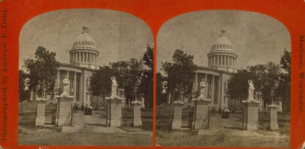 Small group in front of the Wisconsin State Capitol viewed from East Washington Avenue.  Some telegraph wires are strung across foreground. The card is from the series "The Beauties of the City of Madison and Vicinity." The iron and stone fence around the perimeter of the Capitol Park was designed by Stephen V. Shipman, who also designed the Capitol dome, and it was completed in 1873.