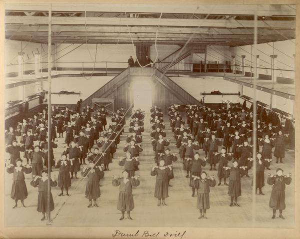 Female students performing a dumbbell drill inside the gymnasium at the Indian Industrial School.