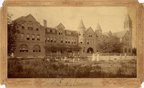 A Cherokee Female Seminary. Students stand on the lawn and porch in front of the building.