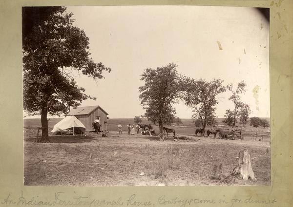 Cowboys come in for dinner at an Indian Territory ranch house. Their horses wait near trees.