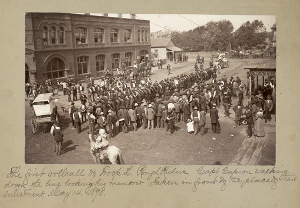 The first roll call of Troop L, Rough Riders. Captain Capron walks down the line, looking his men over. A crowd gathers to watch. The photograph was taken in front of the place of their enlistment.
