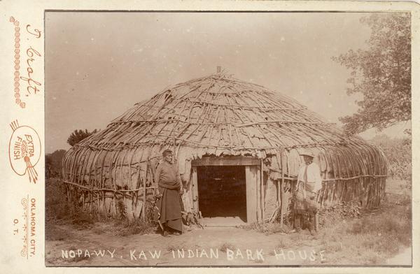 An Indian man and woman stand outside the entrance to a Kaw bark house.