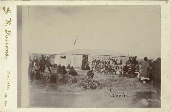 A group of Osage gather in front of a structure to watch a smoke dance.