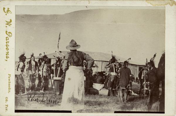 Osage gather in front of a wooden structure for a war dance.