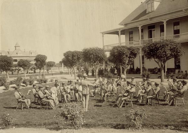 A Pima Agency band performs outside for an audience, which gathers on a nearby porch.
