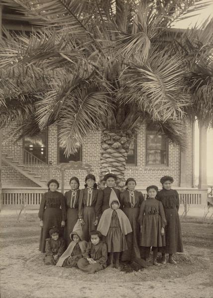 Pima Agency female students and young girls gather in front of a tree on agency grounds.