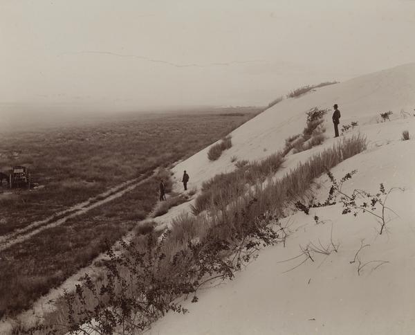 Elevated view from hill of the road between the railroad and the Mescalero Agency, which is surrounded by a sixty by twenty mile section of white sands, composed of pure gypsum, the only formation of its kind in the world. Three men stand on the sands, and on the far left is a wagon.