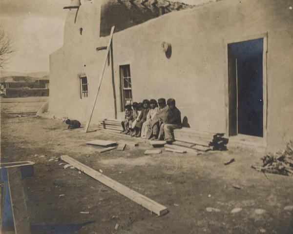 A San Ildefonso Pueblo group sits near a building.