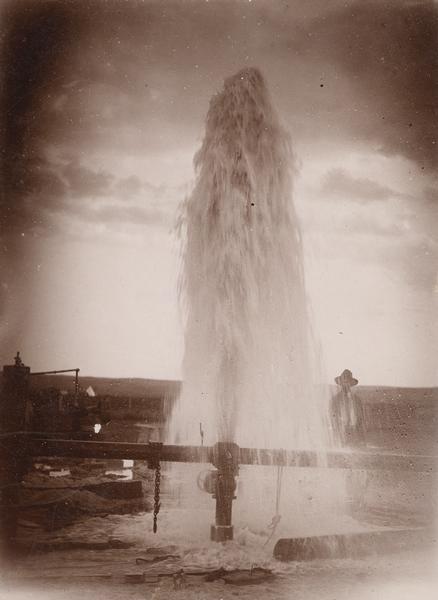 Water gushes 18 feet in the air from a 6-inch pipe at the Lower Brule Agency. A man stands behind the pipe on the right.