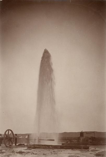 Water gushes 60 feet in the air from a 4-inch pipe at the Lower Brule Agency. Two men stand on the left near pumping machinery.