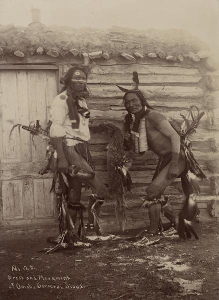 Omaha Sioux Dancers at the Rosebud Agency. The two men are standing in front of a log cabin with a sod roof and wooden door.