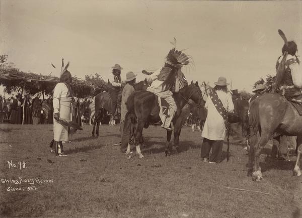 Sioux give away horses at the Rosebud Agency. A large group of people are watching from a shelter on the left covered with branches.