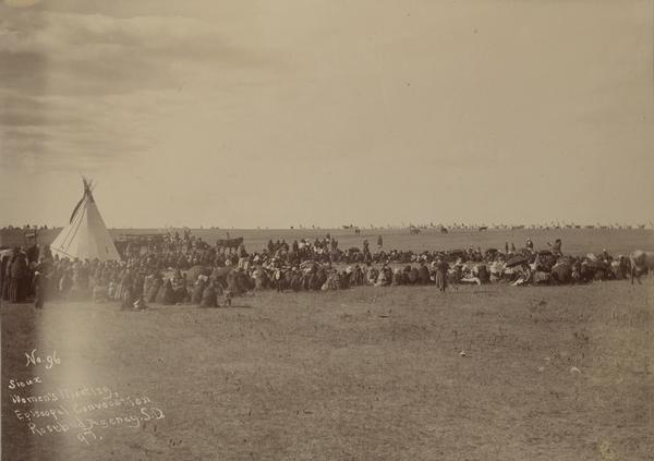 Slightly elevated view across field of a Sioux women's Episcopal Convocation meeting at the Rosebud Agency. The women, and a number of children, are sitting and standing on the open ground. In the background are horses and many tipi's.
