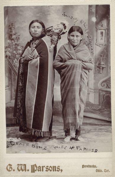 Full-length studio portrait in front of a painted backdrop of Black Bow's wife, with infant in a papoose, and Saucy Chief's squaw.