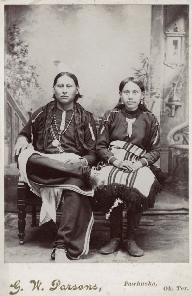 Studio portrait in front of a painted backdrop of an Osage man and woman sitting together.