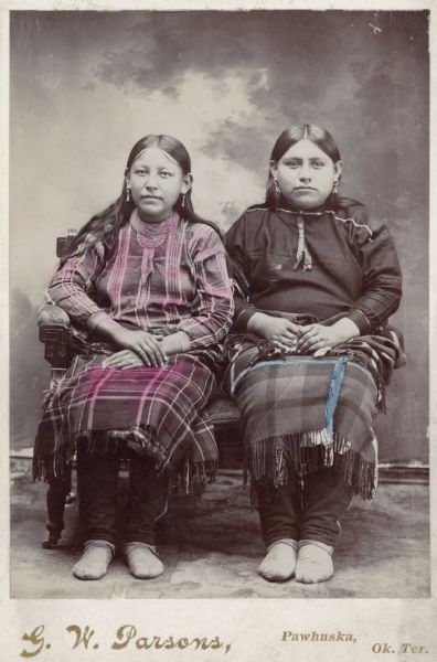 Full-length studio portrait in front of a painted backdrop of two Osage women sitting.