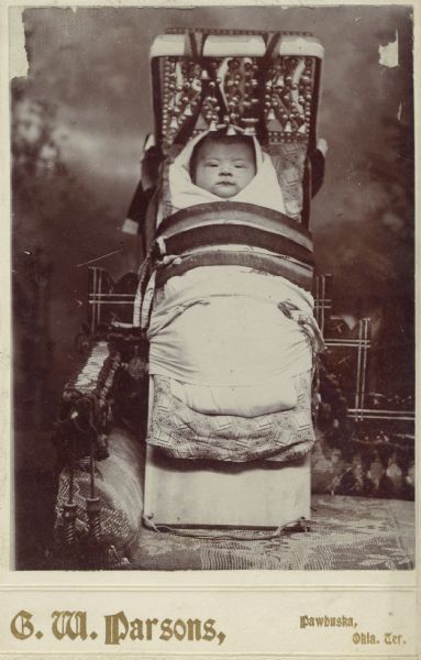 Studio portrait in front of a painted backdrop of an Osage infant, wrapped up in an infant carrier that is resting on a chair.