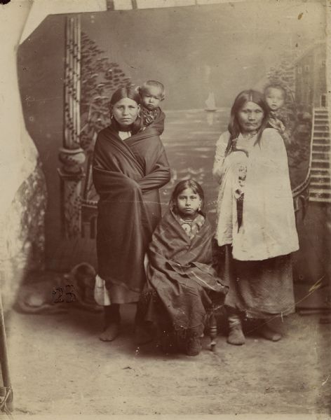 Studio portrait in front of a painted backdrop of two Osage women holding children on their backs in infant carriers, with a young girl sitting between them.