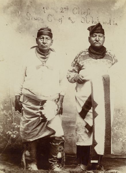 Full-length portrait posed in front of a painted backdrop of the first and second chiefs, Saucy Chief and Olohawalla. This image comes from an Osage album with the inscription: "Compliments of G.W. Parsons."