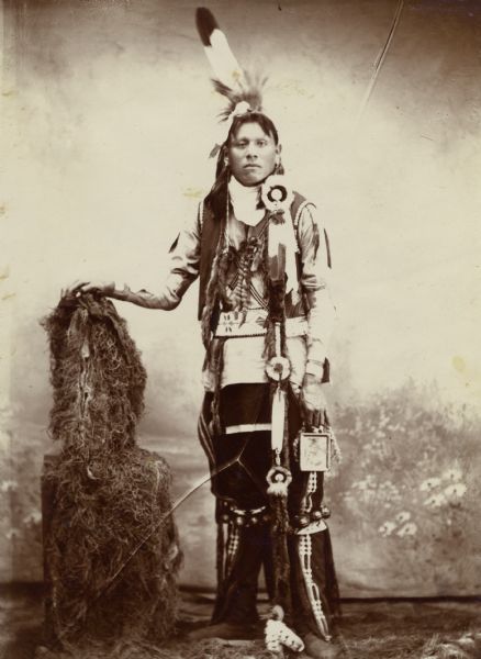Full-length studio portrait in front of a painted backdrop of an Osage man in full dress. This image comes from an Osage album with the inscription: "Compliments of G.W. Parsons."