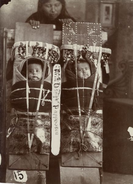 A woman stands behind twin Osage papooses in a studio portrait in front of a painted backdrop. The infants are tightly wrapped up. This image comes from an Osage album with the inscription: "Compliments of G.W. Parsons."