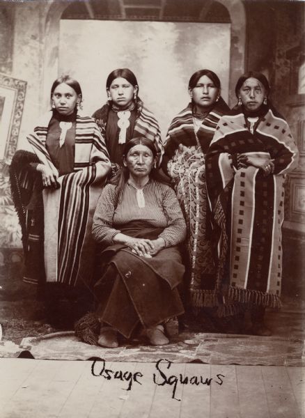 Five "Osage squaws" gather for a portrait. They are posed in front of a painted backdrop. This image comes from an Osage album with the inscription: "Compliments of G.W. Parsons."