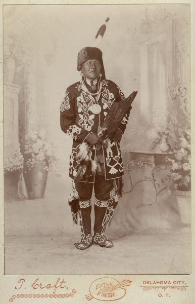 Full-length portrait of White Horse, an Otoe chief, wearing the clothing of the Revitalization Movement, and posing in front of a painted backdrop.