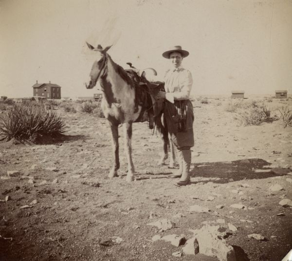 A woman, possibly Sarah Jones, wife of William Arthur Jones, with a horse. In the far background is a house and other buildings.